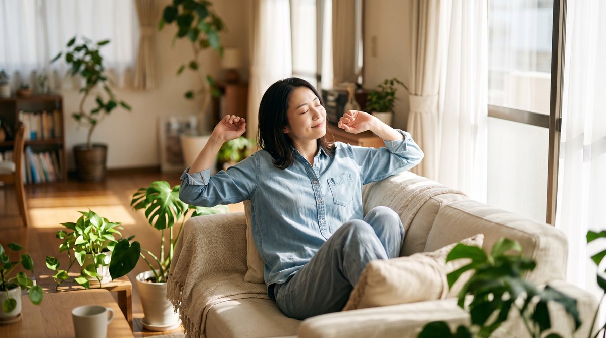 休日の朝、日の当たる窓辺のソファでのびをする女性